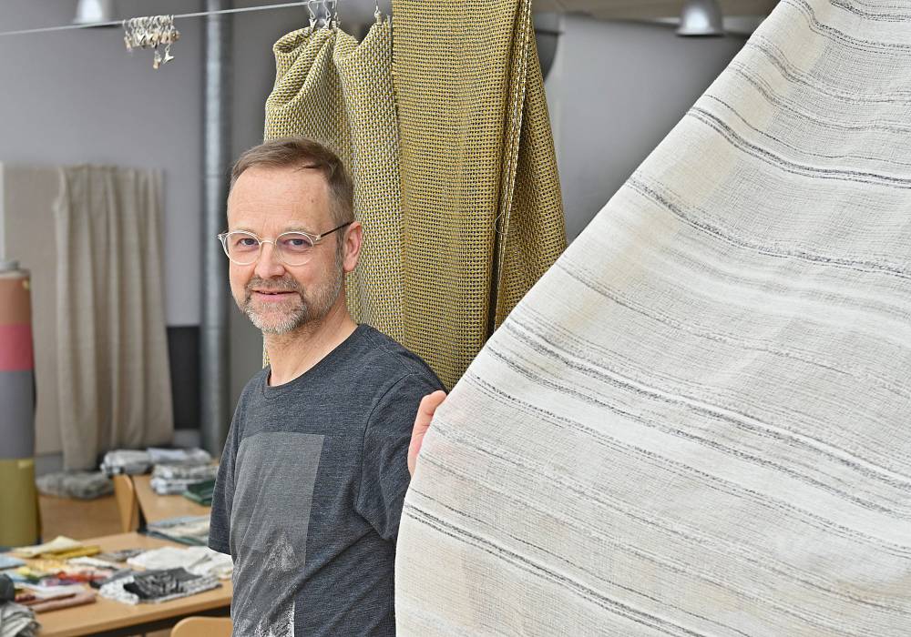 Textildesigner präsentiert großen Möbelstoff in Atelier mit hängenden Stoffmustern.